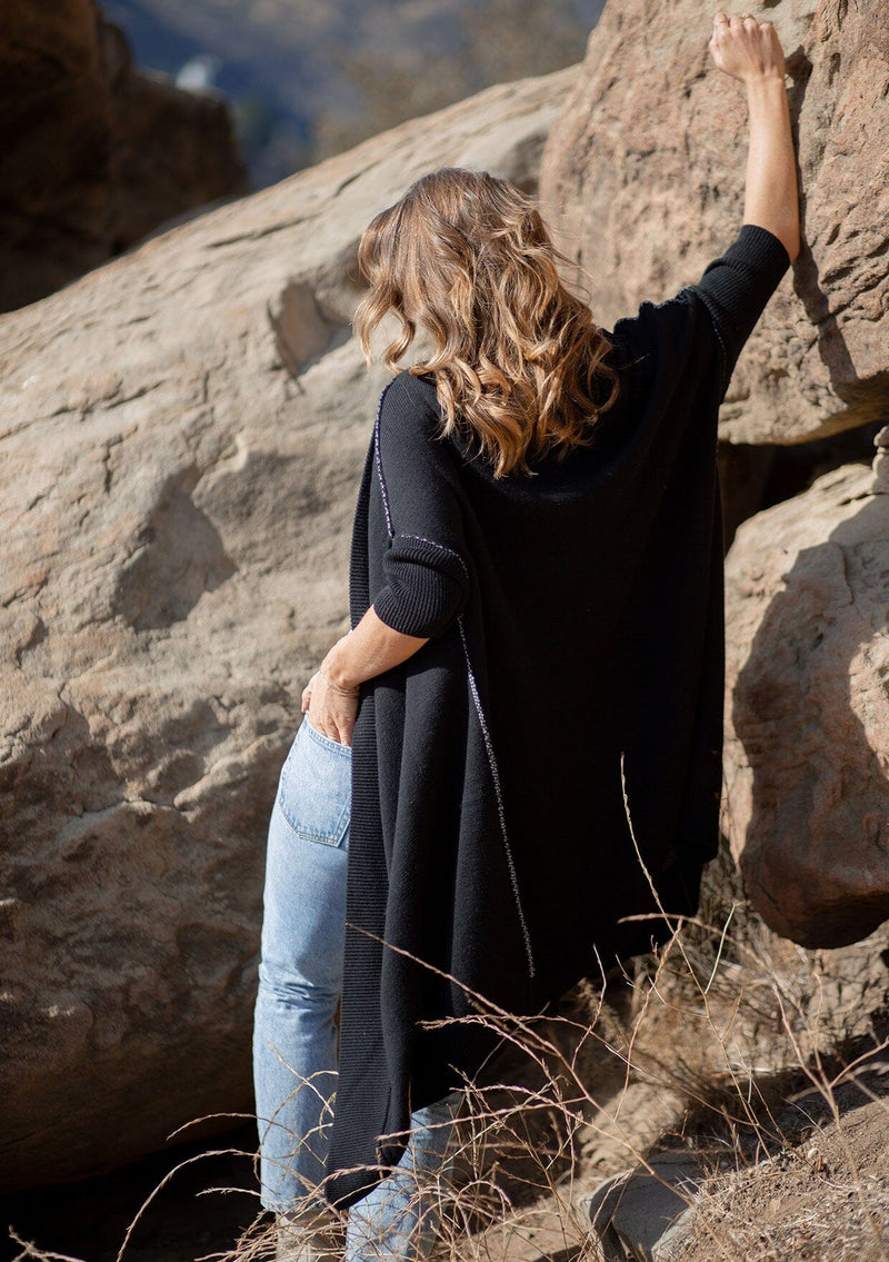 [Color: Black] A blonde model wearing a fall black cardigan with a contrast interior, exposed seam details, and a tapered sleeve at the elbow. 