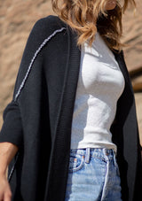 [Color: Black] A blonde model wearing a fall black cardigan with a contrast interior, exposed seam details, and a tapered sleeve at the elbow. 