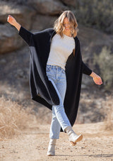 [Color: Black] A blonde model wearing a fall black cardigan with a contrast interior, exposed seam details, and a tapered sleeve at the elbow. 