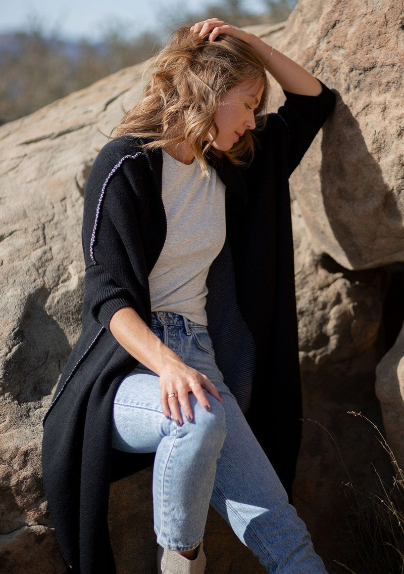 [Color: Black] A blonde model wearing a fall black cardigan with a contrast interior, exposed seam details, and a tapered sleeve at the elbow. 