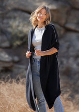 [Color: Black] A blonde model wearing a fall black cardigan with a contrast interior, exposed seam details, and a tapered sleeve at the elbow. 