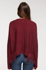 [Color: Merlot] A back facing image of a brunette model wearing a burgundy knit pullover with side fringe details, long sleeves with dropped shoulders, and a round neckline. 