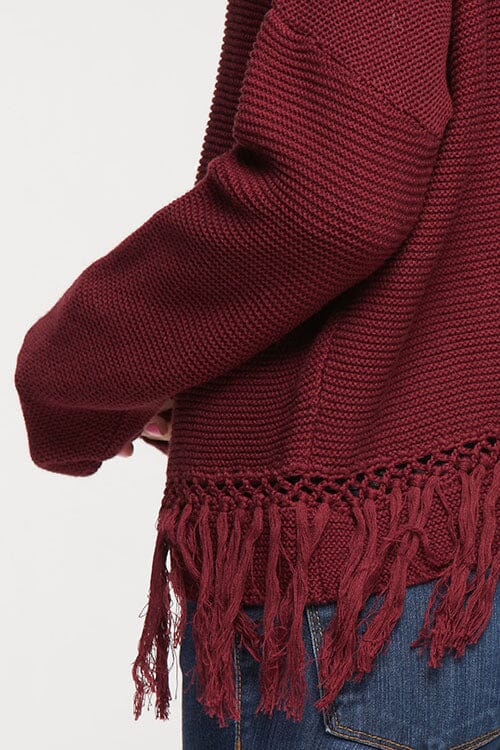[Color: Merlot] A detail image of a brunette model wearing a burgundy knit pullover with side fringe details, long sleeves with dropped shoulders, and a round neckline. 