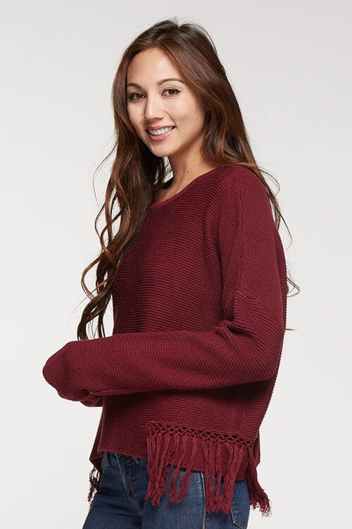[Color: Merlot] A side facing image of a brunette model wearing a burgundy knit pullover with side fringe details, long sleeves with dropped shoulders, and a round neckline. 