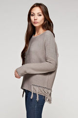 [Color: Mushroom] A side facing image of a brunette model wearing a grey knit pullover with side fringe details, long sleeves with dropped shoulders, and a round neckline. 