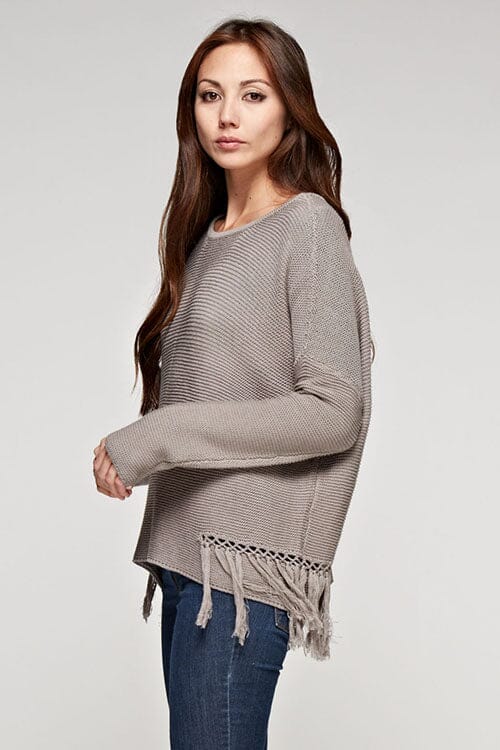 [Color: Mushroom] A side facing image of a brunette model wearing a grey knit pullover with side fringe details, long sleeves with dropped shoulders, and a round neckline. 