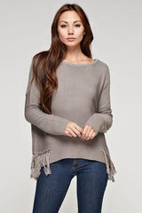 [Color: Mushroom] A front facing image of a brunette model wearing a grey knit pullover with side fringe details, long sleeves with dropped shoulders, and a round neckline. 
