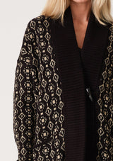 [Color: Black/Taupe] A close up front facing image of a blonde model wearing a cozy bohemian cardigan in a black and taupe geo jacquard design. With contrast ribbed trim, side pockets, a shawl collar, and a toggle button closure. 
