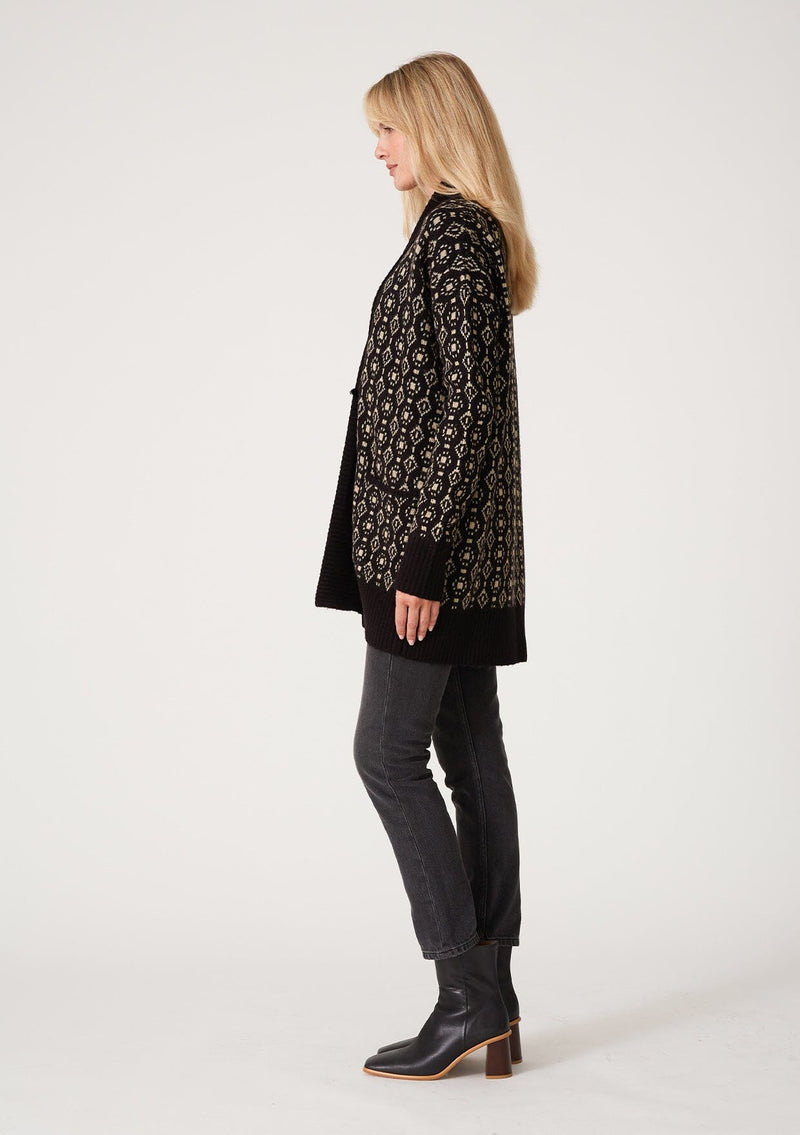 [Color: Black/Taupe] A side facing image of a blonde model wearing a cozy bohemian cardigan in a black and taupe geo jacquard design. With contrast ribbed trim, side pockets, a shawl collar, and a toggle button closure. 