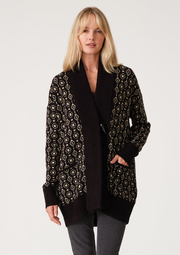 [Color: Black/Taupe] A front facing image of a blonde model wearing a cozy bohemian cardigan in a black and taupe geo jacquard design. With contrast ribbed trim, side pockets, a shawl collar, and a toggle button closure. 