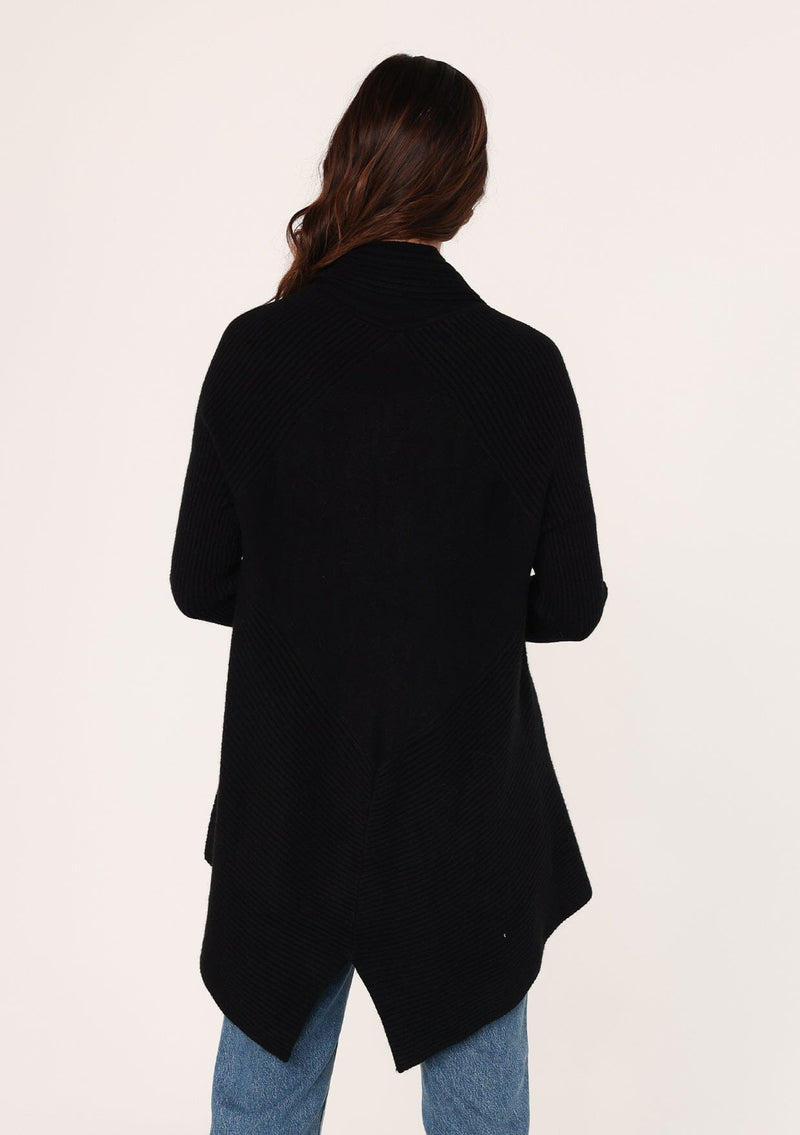 [Color: Black] A back facing image of a brunette model wearing a black shawl cardigan with a ribbed texture, open front, and a back vent detail. Cozy and relaxed for the fall season. 
