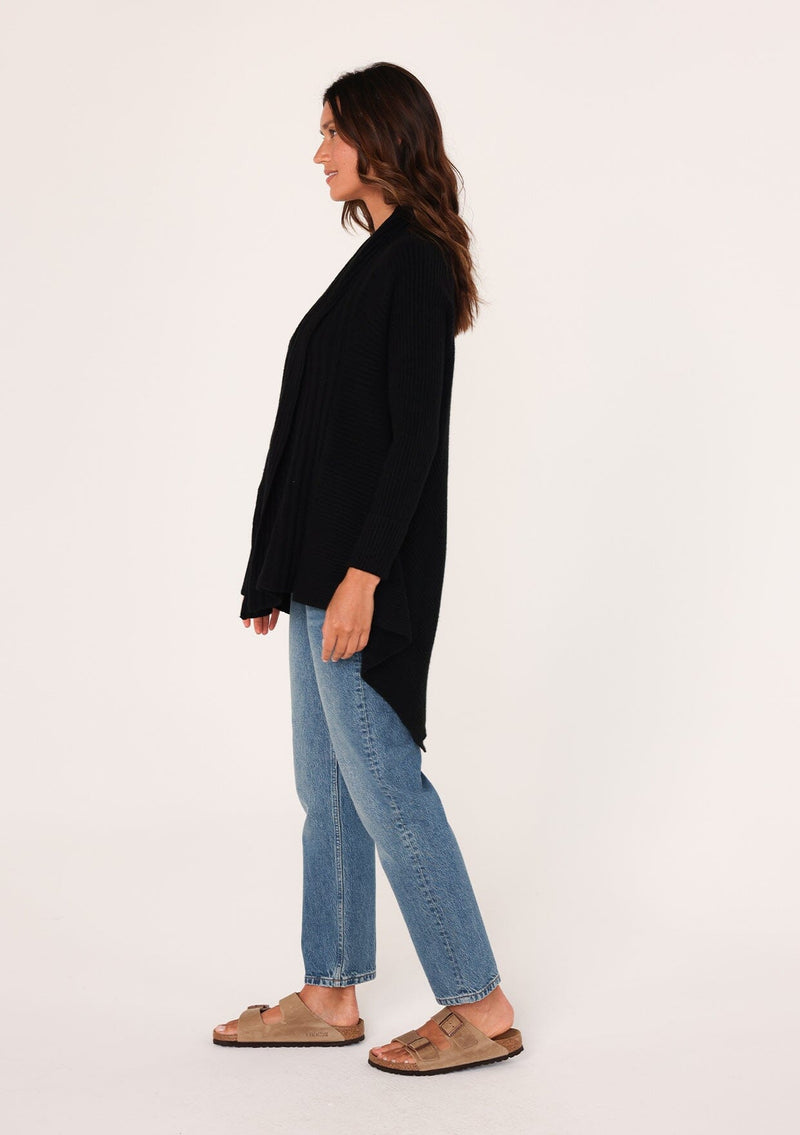 [Color: Black] A side facing image of a brunette model wearing a black shawl cardigan with a ribbed texture, open front, and a back vent detail. Cozy and relaxed for the fall season. 