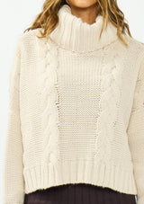 [Color: Bone] A detail image of a brunette model wearing a cream cable knit sweater with a turtleneck over a brown maxi skirt. 