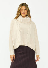 [Color: Bone] A front facing image of a brunette model wearing a cream cable knit sweater with a turtleneck over a brown maxi skirt. 