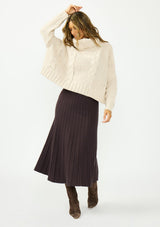 [Color: Bone] A front facing image of a brunette model wearing a cream cable knit sweater with a turtleneck over a brown maxi skirt. 