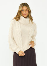[Color: Bone] A front facing image of a brunette model wearing a cream cable knit sweater with a turtleneck over a brown maxi skirt. 
