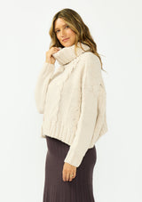 [Color: Bone] A side facing image of a brunette model wearing a cream cable knit sweater with a turtleneck over a brown maxi skirt. 
