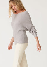 [Color: Light Heather Grey] A side facing image of a blonde model wearing a light heather grey waffle knit pullover sweater. With long sleeves, a relaxed fit, and a wide neckline that can be worn off the shoulder.