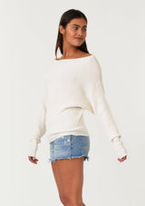 [Color: Natural] A side facing image of a brunette model wearing an off white waffle knit pullover sweater. With long sleeves, a relaxed fit, and a wide neckline that can be worn off the shoulder.