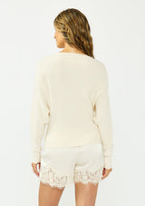 [Color: Natural] A back facing image of a brunette model wearing an off white pullover sweater with a boat neckline and long sleeves that taper at the cuffs. Styled with off white satin lace shorts. 