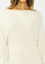 [Color: Natural] A detail image of a brunette model wearing an off white pullover sweater with a boat neckline and long sleeves that taper at the cuffs. Styled with off white satin lace shorts. 