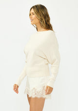 [Color: Natural] A side facing image of a brunette model wearing an off white pullover sweater with a boat neckline and long sleeves that taper at the cuffs. Styled with off white satin lace shorts. 