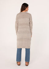 [Color: Oatmeal] A back view of a brunette woman modeling a long, open-front beige mélange knit cardigan over a white top and blue jeans.