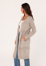 [Color: Oatmeal] A side view of a brunette woman modeling a long, open-front beige mélange knit cardigan over a white top and blue jeans.
