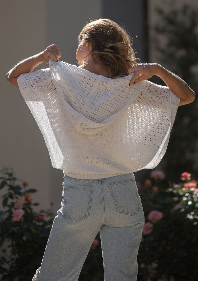[Color: Heather Off White] A back facing image of a blonde model wearing a pointelle knit sweater top with a surplice v-neckline, short dolman sleeves,  wide sleeve openings, and fitted ribbed hem. 