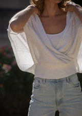 [Color: Heather Off White] A detail image of a blonde model wearing a pointelle knit sweater top with a surplice v-neckline, short dolman sleeves,  wide sleeve openings, and fitted ribbed hem. 