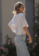 [Color: Heather Off White] A side facing image of a blonde model wearing a pointelle knit sweater top with a surplice v-neckline, short dolman sleeves,  wide sleeve openings, and fitted ribbed hem. 