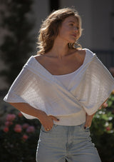 [Color: Heather Off White] A front facing image of a blonde model wearing a pointelle knit sweater top with a surplice v-neckline, short dolman sleeves,  wide sleeve openings, and fitted ribbed hem. 