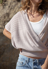[Color: Heather Taupe] A detail image of a blonde model wearing a pointelle knit sweater top with a surplice v-neckline, short dolman sleeves,  wide sleeve openings, and fitted ribbed hem. 