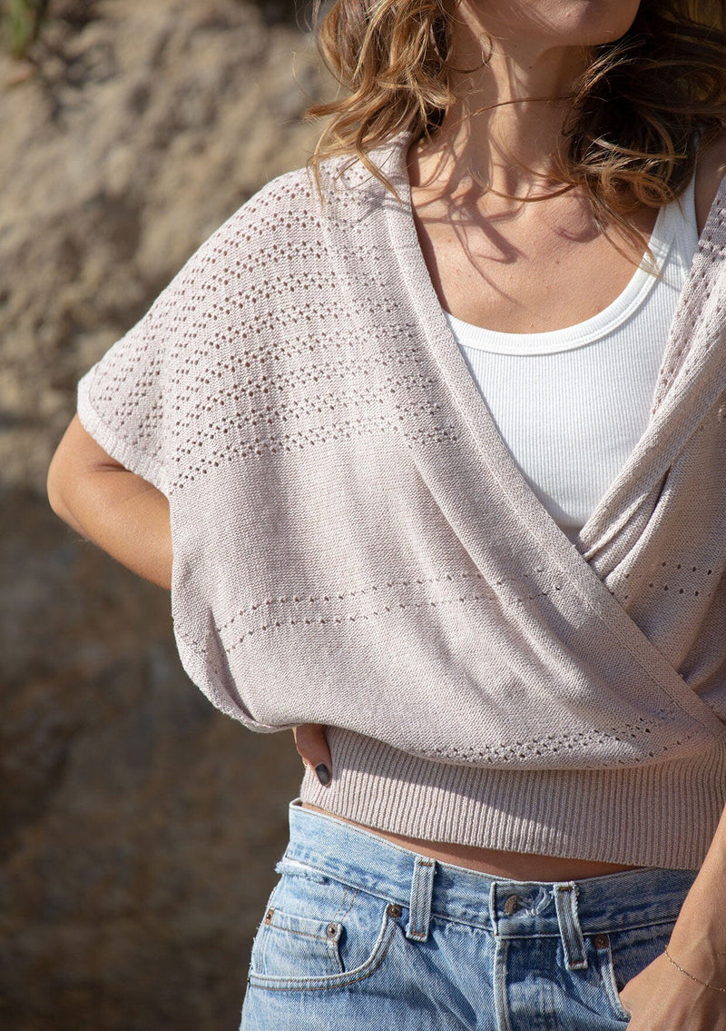 [Color: Heather Taupe] A detail image of a blonde model wearing a pointelle knit sweater top with a surplice v-neckline, short dolman sleeves,  wide sleeve openings, and fitted ribbed hem. 