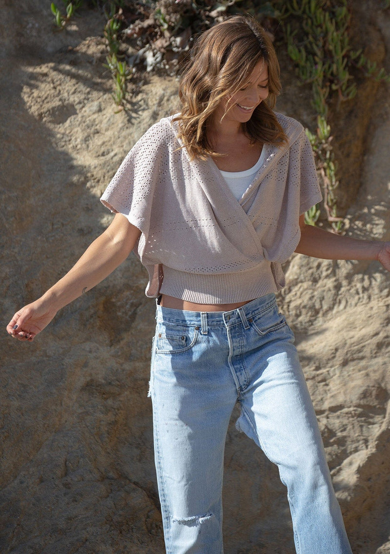 [Color: Heather Taupe] A front facing image of a blonde model wearing a pointelle knit sweater top with a surplice v-neckline, short dolman sleeves,  wide sleeve openings, and fitted ribbed hem. 