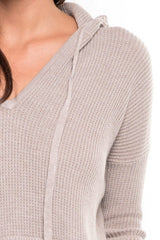 [Color: Heather Cement] A detail image of a brunette model wearing a taupe waffle knit pullover with a drawstring hoodie, kangaroo pockets, and side slits with tie detail. 