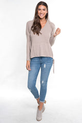 [Color: Heather Cement] A front facing image of a brunette model wearing a taupe waffle knit pullover with a drawstring hoodie, kangaroo pockets, and side slits with tie detail. 