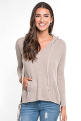 [Color: Heather Cement] A front facing image of a brunette model wearing a taupe waffle knit pullover with a drawstring hoodie, kangaroo pockets, and side slits with tie detail. 