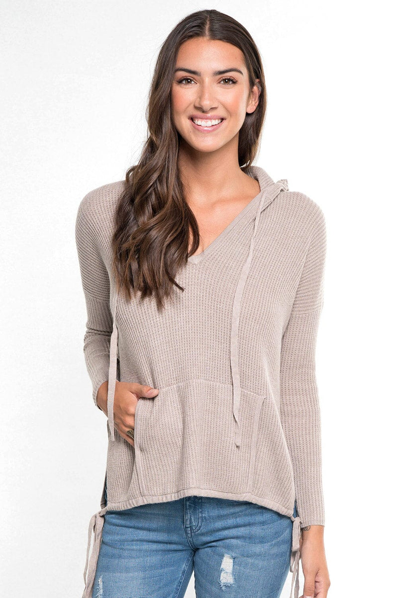 [Color: Heather Cement] A front facing image of a brunette model wearing a taupe waffle knit pullover with a drawstring hoodie, kangaroo pockets, and side slits with tie detail. 
