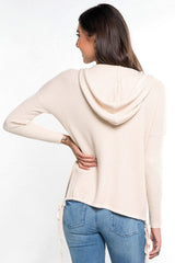 [Color: Vanilla] A back facing image of a brunette model wearing a cream waffle knit pullover with a drawstring hoodie, kangaroo pockets, and side slits with tie detail. 