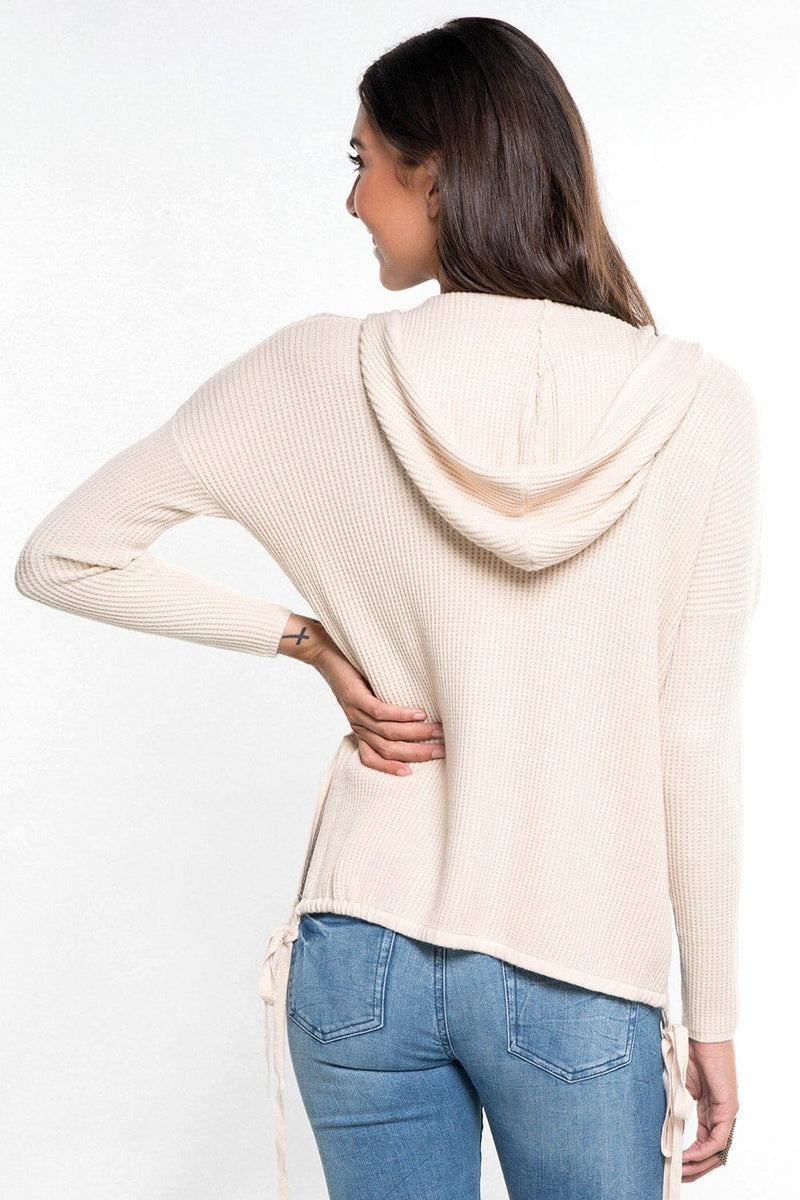 [Color: Vanilla] A back facing image of a brunette model wearing a cream waffle knit pullover with a drawstring hoodie, kangaroo pockets, and side slits with tie detail. 