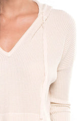 [Color: Vanilla] A detail image of a brunette model wearing a cream waffle knit pullover with a drawstring hoodie, kangaroo pockets, and side slits with tie detail. 