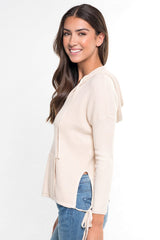 [Color: Vanilla] A side facing image of a brunette model wearing a cream waffle knit pullover with a drawstring hoodie, kangaroo pockets, and side slits with tie detail. 