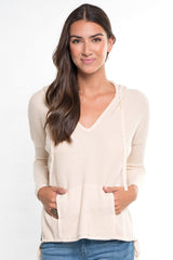 [Color: Vanilla] A front facing image of a brunette model wearing a cream waffle knit pullover with a drawstring hoodie, kangaroo pockets, and side slits with tie detail. 