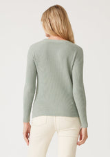 [Color: Dusty Sage] A back facing image of a blonde model wearing a dusty sage green waffle knit pullover sweater. With long sleeves, a wide crew neckline, and a twist front detail at the waist. 