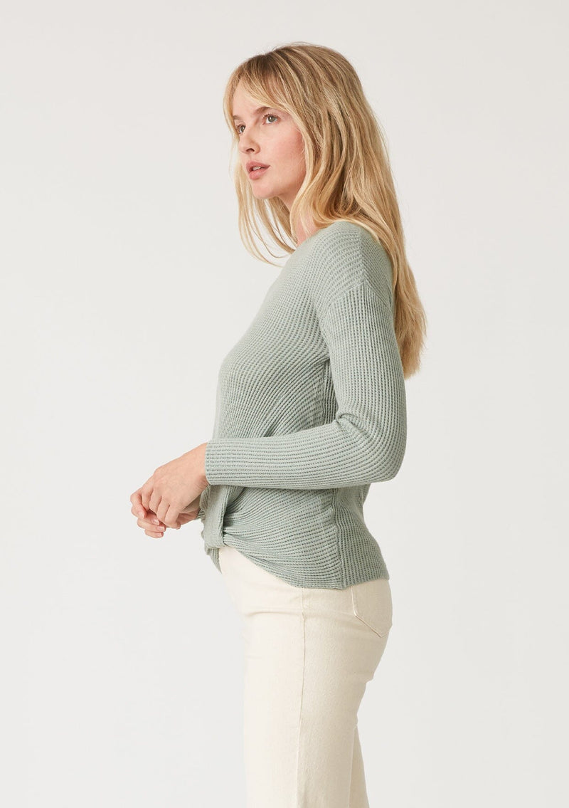 [Color: Dusty Sage] A side facing image of a blonde model wearing a dusty sage green waffle knit pullover sweater. With long sleeves, a wide crew neckline, and a twist front detail at the waist. 
