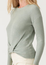 [Color: Dusty Sage] A close up side facing image of a blonde model wearing a dusty sage green waffle knit pullover sweater. With long sleeves, a wide crew neckline, and a twist front detail at the waist. 