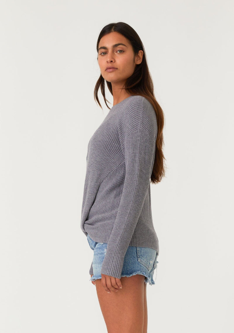 [Color: Heather Charcoal] A side facing image of a brunette model wearing a heather grey waffle knit pullover sweater. With long sleeves, a wide crew neckline, and a twist front detail at the waist.