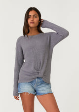 [Color: Heather Charcoal] A front facing image of a brunette model wearing a heather grey waffle knit pullover sweater. With long sleeves, a wide crew neckline, and a twist front detail at the waist.