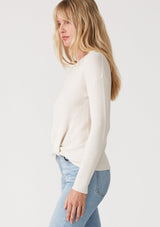 [Color: Natural] A side facing image of a blonde model wearing an ivory waffle knit pullover sweater. With long sleeves, a wide crew neckline, and a twist front detail at the waist. 